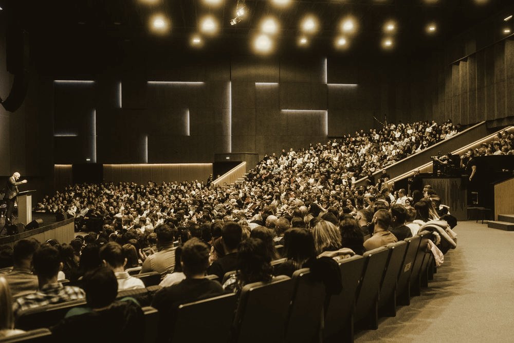 A packed auditorium audience facing a stage under blue conference lighting